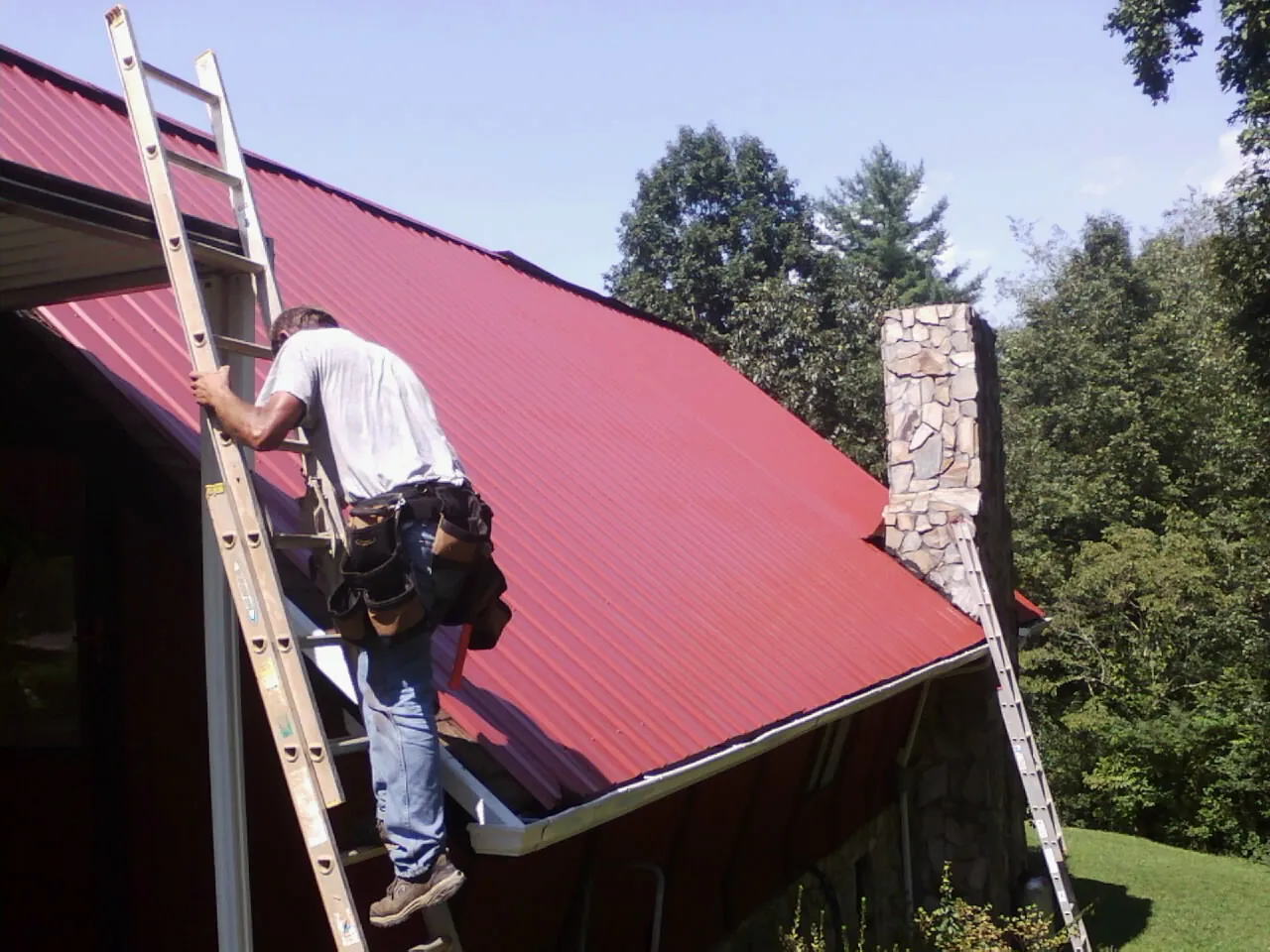 Roofer on a ladder beside a red metal roof and stone chimney — Soffit & Fascia Repair by Roofing Brothers in Hadley